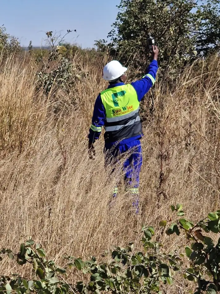 SunWise technician in branded PPE holding a handheld air quality meter above dry grassland