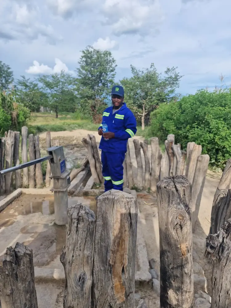 SunWise technician standing beside a fenced community hand-pump borehole holding a sample bottle