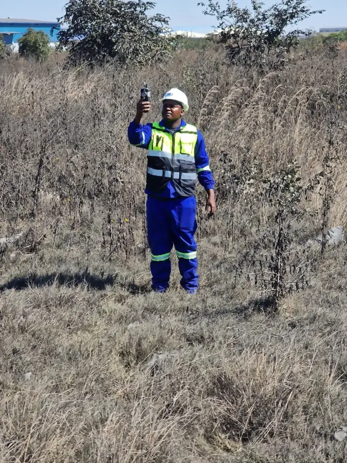 SunWise technician holding a handheld air quality meter overhead in dry grassland, industrial buildings in the distance