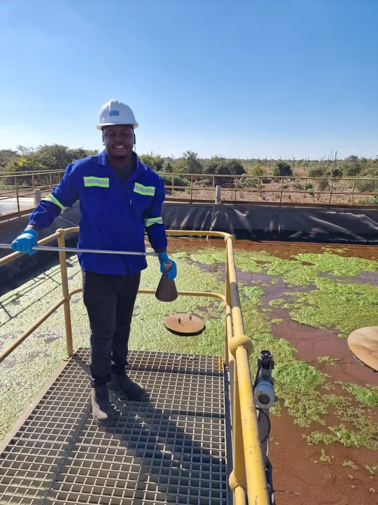 SunWise technician in gloves holding a sampling dipper on the grating walkway above a lined effluent pond