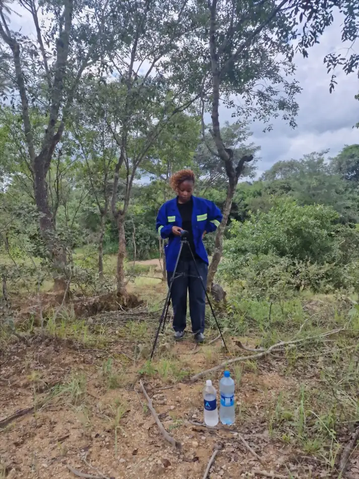 SunWise technician standing behind a tripod-mounted monitoring instrument in woodland, sample bottles on the ground